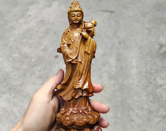 May include: A wooden statue of a female figure with a crown and a lotus flower in her hand. The statue is standing on a lotus base and is painted in shades of brown.
