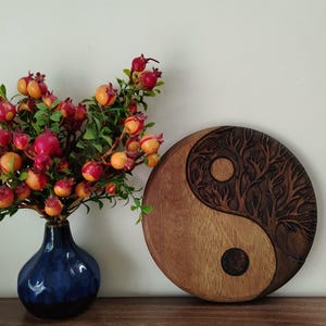 May include: A wooden Yin and Yang wall hanging with a tree design carved into one side. The other side is smooth. A dark blue vase with red and orange berries sits to the left.