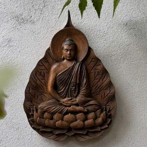 Könnte beinhalten: Braune Holzstatue eines sitzenden Buddha in meditativer Haltung, in Relief geschnitzt. Die Statue befindet sich vor einer strukturierten weißen Wand, mit grünen Blättern im Hintergrund. Der Buddha sitzt auf einer Lotusblume.