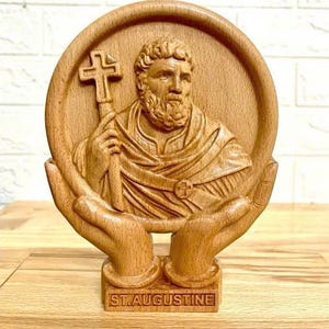 Op de afbeelding: Houten beeldhouwwerk van St. Augustinus, met een gedetailleerde afbeelding van de heilige die een staf met een kruis vasthoudt. De gravure is geplaatst in een cirkelvormig frame, ondersteund door gebeeldhouwde handen, en bevat de tekst "ST. AUGUSTINE" aan de basis.