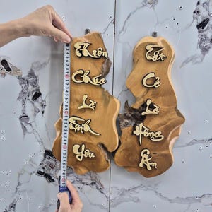 以下が含まれることがあります： ベトナム語の文字が書かれた2つの木製の壁掛け。木材は温かみのある茶色で、文字は対照的な金色です。壁掛けの高さは約76cmです。