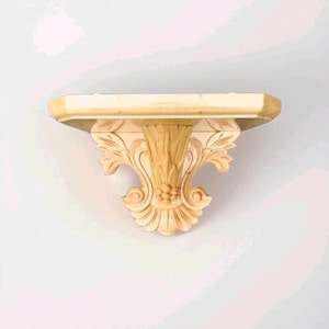 May include: A light-coloured wooden wall shelf featuring a flat rectangular top surface supported by a decorative bracket. The bracket displays intricate carved leaf-like patterns and a central vertical fluted design with a small cluster of spheres.