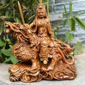 May include: A detailed wooden sculpture of a seated figure holding a sword, riding a dragon. The sculpture is carved from light brown wood, with intricate details in the dragon's scales and the figure's clothing. The background features green foliage.