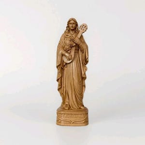 Op de afbeelding: Een lichtbruin houten standbeeld van de heilige Cecilia op een rechthoekige voet. De figuur heeft lang haar, draagt gedrapeerde gewaden en houdt een snaarinstrument vast met rozen in de kleding. De naam ST. CECILIA is in de voorzijde van de voet gegraveerd.