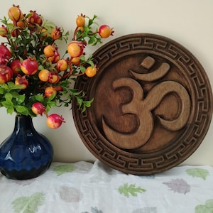 Peut inclure: Décoration murale ronde en bois avec le symbole Om sculpté au centre, entouré d'une bordure décorative. Un vase bleu contient un bouquet de baies rouges et jaunes à gauche.