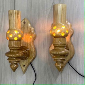 May include: A pair of wooden wall sconces in a light brown hue. Each features a carved design with a rounded central section, illuminated by a warm yellow light. They are mounted on decorative wooden backplates, with a cable attached.