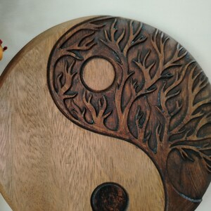 May include: A wooden Yin and Yang symbol with a carved tree design. The symbol is split into two sections, one light brown and the other dark brown with a tree carving. A circle is carved into each section.