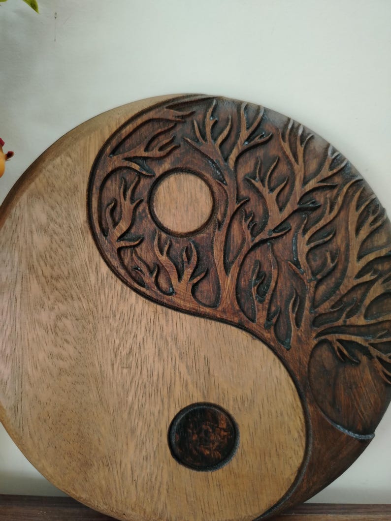 May include: A wooden Yin and Yang symbol with a carved tree design. The symbol is split into two sections, one light brown and the other dark brown with a tree carving. A circle is carved into each section.