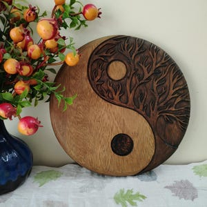 May include: A round wooden Yin and Yang wall hanging. One side is a lighter wood, the other is a darker wood with a carved tree design. The piece is displayed on a white cloth with green leaf patterns, next to a blue vase with red and orange berries.
