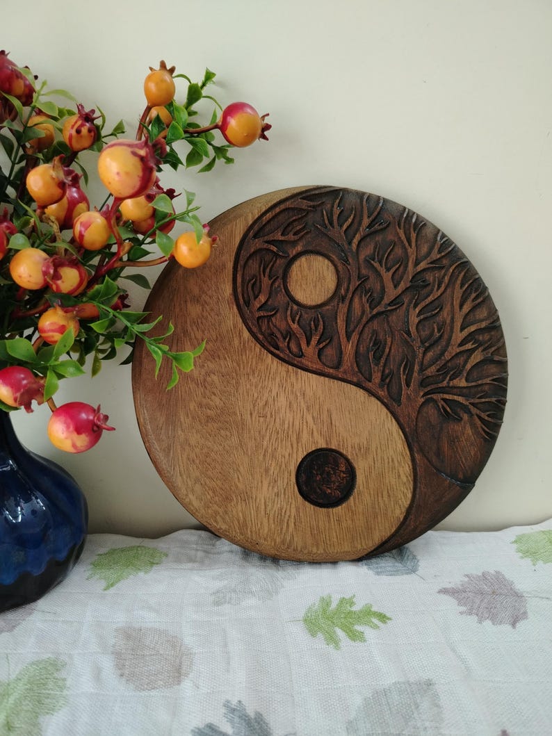 May include: A round wooden Yin and Yang wall hanging. One side is a lighter wood, the other is a darker wood with a carved tree design. The piece is displayed on a white cloth with green leaf patterns, next to a blue vase with red and orange berries.