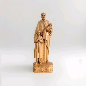 May include: A light brown wooden figurine depicting Saint Vincent de Paul standing while holding a small child in one arm, with a young child standing beside him with hands clasped in prayer. The base features the text St. Vincent de Paul.