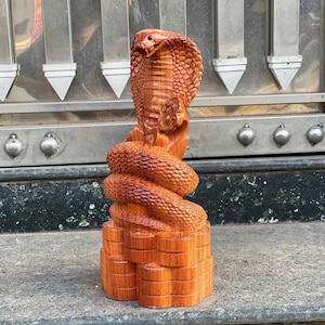 May include: A detailed wooden sculpture of a cobra snake. The snake is coiled, with its hood raised, and is carved from a reddish-brown wood. The sculpture is set on a tiered base, showcasing the intricate details of the carving.