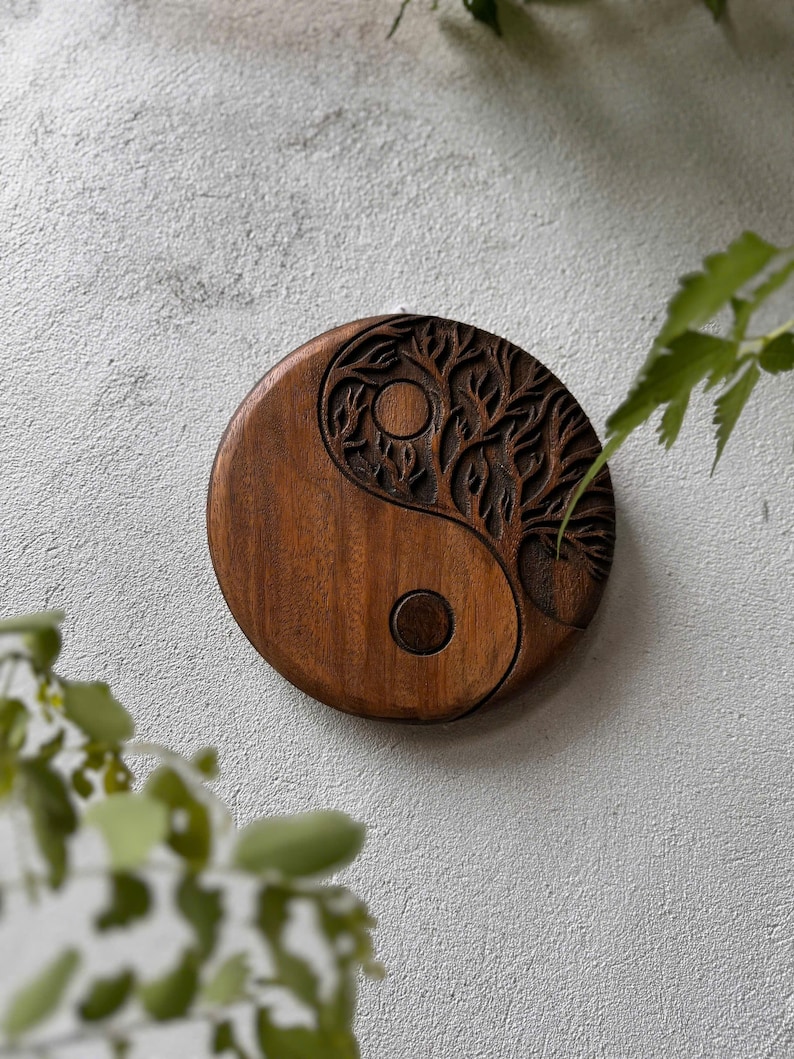 May include: A round, wooden Yin and Yang symbol with a tree of life design carved into one half. The wood is a warm brown color, and the carving is detailed. The symbol is set against a textured, light gray background.