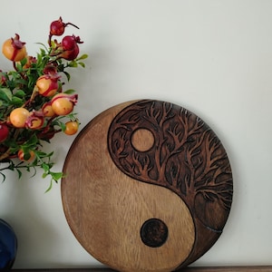 May include: A round wooden Yin and Yang symbol with a tree carved into one half. The other half is smooth. The symbol is brown with a lighter brown half. A vase of flowers is in the background.