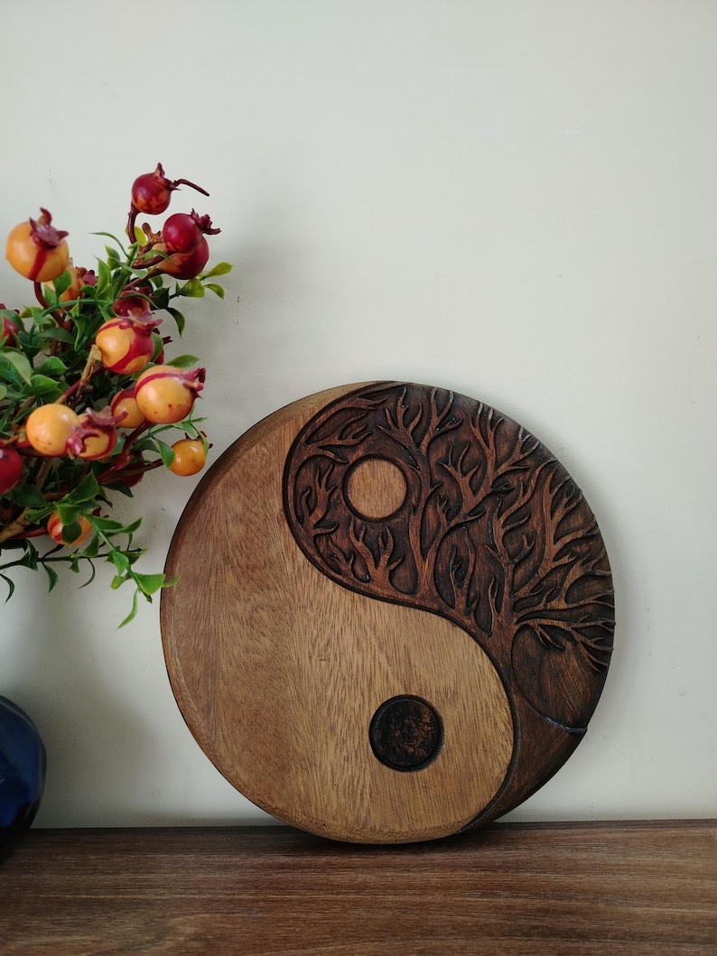 May include: A round wooden Yin and Yang symbol with a tree carved into one half. The other half is smooth. The symbol is brown with a lighter brown half. A vase of flowers is in the background.