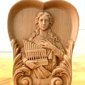 Op de afbeelding: Een houten reliëfsculptuur van Sint-Cecilia, met een kleine orgel. De sculptuur staat in een hartvormige lijst met sierlijke krullen en de tekst "St. Cecilia" onderaan. Het hout heeft een lichtbruine kleur.