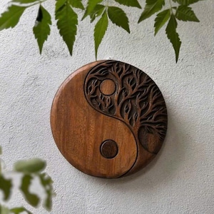 May include: A round, wooden Yin and Yang wall hanging. One side features a carved tree design, the other a circle. The wood is a warm brown color. Green leaves are in the background.