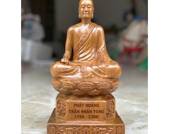 May include: A wooden statue of a seated figure in a meditative pose. The figure is wearing a robe and is seated on a lotus flower base. The base has engraved text. The statue is made of light brown wood.