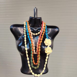 May include: A black mannequin displays several colorful crochet necklaces. The necklaces include white, orange, blue, green, and yellow strands. Two yellow crochet flower accents are pinned to the mannequin.