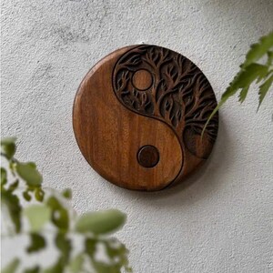 May include: A round, wooden wall hanging featuring a carved Yin and Yang symbol. The dark brown wood is intricately carved with a tree design on one side and a circular design on the other. The wall hanging is displayed against a textured, light gray wall.