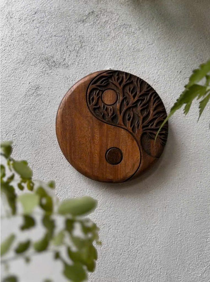 May include: A round, wooden wall hanging featuring a carved Yin and Yang symbol. The dark brown wood is intricately carved with a tree design on one side and a circular design on the other. The wall hanging is displayed against a textured, light gray wall.