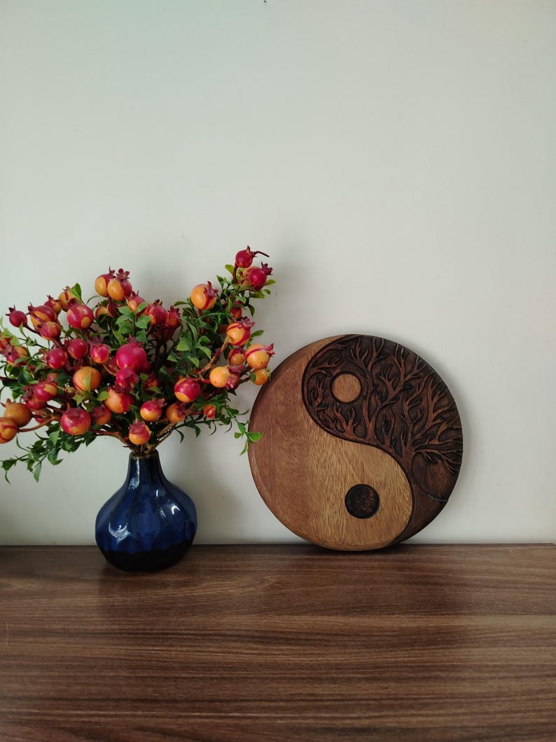 May include: A wooden Yin and Yang wall hanging with a tree design carved into one side. The other side is smooth. A dark blue vase with red and orange berries sits to the left.