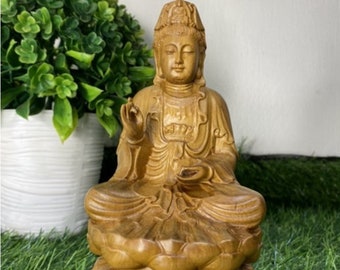 May include: A wooden statue of a seated Buddha, carved in a light brown color. The Buddha is wearing a robe and has a serene expression on their face. The statue is sitting on a lotus flower base.