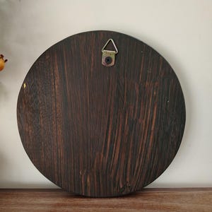 May include: A round, dark brown wooden plaque with a visible wood grain pattern. A metal hanger is attached to the top center for wall mounting. The plaque is displayed on a wooden surface.