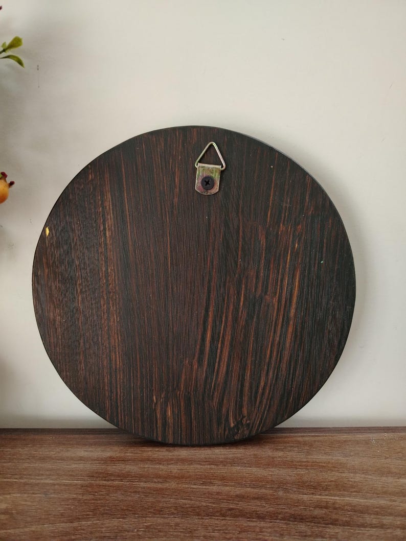 May include: A round, dark brown wooden plaque with a visible wood grain pattern. A metal hanger is attached to the top center for wall mounting. The plaque is displayed on a wooden surface.