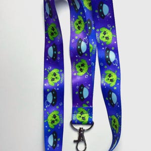 May include: A purple lanyard featuring a repeating pattern of green alien faces and flying saucers. It has a silver metal clasp. The background is dark blue with star shapes.