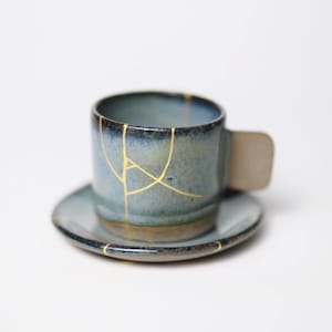 May include: A blue ceramic teacup and saucer set, featuring gold-colored kintsugi repair lines. The cup has a small handle and sits on a matching saucer. The set is a unique example of Japanese pottery.