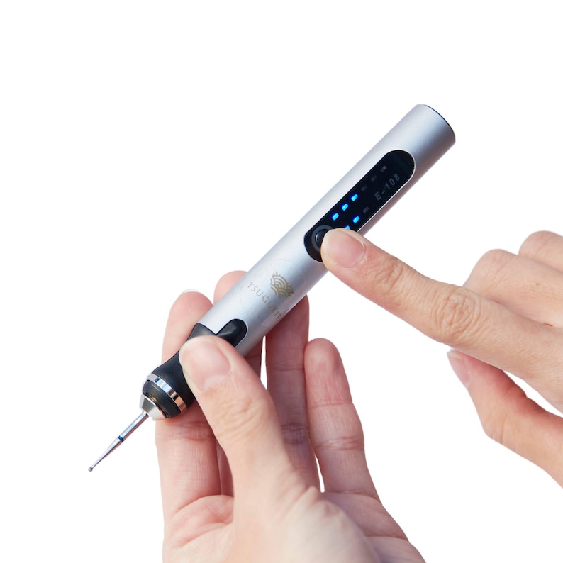 May include: A silver electric nail drill with a blue LED display and a black button. The drill has a small, silver bit attached to the end. The drill is being held in a person's hand.