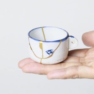 Könnte beinhalten: Kleine weiße Keramiktasse mit blauem Rand und Henkel, verziert mit goldfarbenen Kintsugi-Reparaturlinien. Die Tasse wird in einer Hand vor weißem Hintergrund gehalten. Die Tasse hat einen Durchmesser von ca. 5 cm.