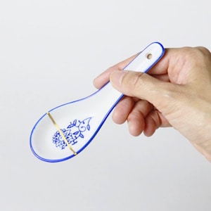 May include: White ceramic spoon with blue floral design and gold accents. The spoon has a blue rim and a small hole in the handle. The spoon is held in a hand.
