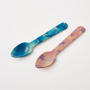 May include: Two wooden spoons with a painted design. One spoon is blue with gold accents, and the other is pink with purple and gold accents. The spoons have an oval bowl and a flat handle.