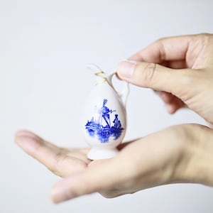 Puede incluir: Una pequeña jarra de cerámica blanca con un diseño azul y dorado. La jarra presenta una ilustración azul de un molino de viento y figuras. El asa y el borde están acentuados con dorado. La jarra está sostenida en las manos de una persona.