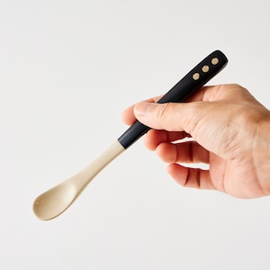 May include: A black and beige spoon with a rounded bowl. The handle is black with three gold-colored dots near the top. The spoon is held in a hand against a white background. The spoon is likely for serving or eating.