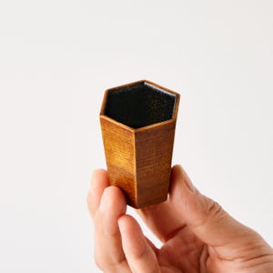May include: A small, hexagonal wooden cup with a dark interior. The cup is held in a hand and has a natural wood grain finish. The interior appears to be coated with a black, glossy material. The cup's shape is tapered, wider at the top and narrower at the base.