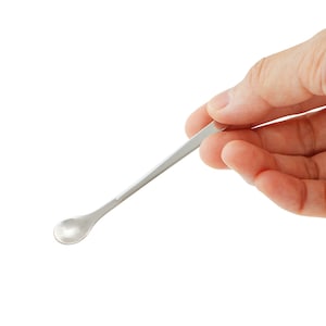 May include: A small, silver laboratory spatula with a rounded spoon-like end and a long, flat handle. The spatula is held in a person's hand against a white background. This tool is used for scooping and transferring small amounts of substances.