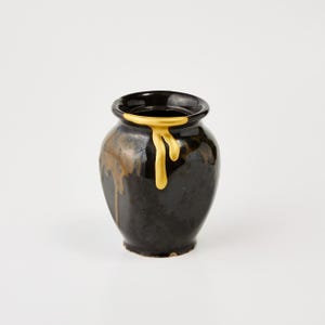 May include: A small, black ceramic vase with a rounded body and a wide, slightly flared rim. The vase has a glossy finish with gold-colored drips cascading down the side, creating a striking contrast. The interior of the vase is also black.