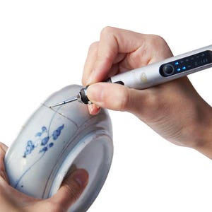 May include: A person is using a small, silver electric tool with a blue light to repair a crack in a white ceramic bowl with a blue and white floral design.