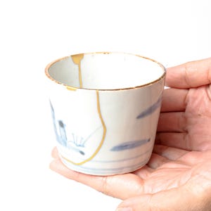 Könnte beinhalten: Ein kleiner weißer Keramikbecher mit blauen Pinselstrichmustern und goldfarbenen Lacklinien, die Risse am Rand und am Körper reparieren. Der Becher hat eine zylindrische Form mit einer glatten Oberfläche und einem goldfarbenen Rand.