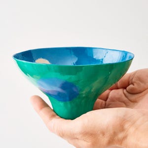 May include: A small, hand-formed bowl with an irregular, organic shape. The exterior is a vibrant green with a large, rounded blue patch, while the interior is finished in a bright, solid blue. The surface has a glossy, reflective glaze.