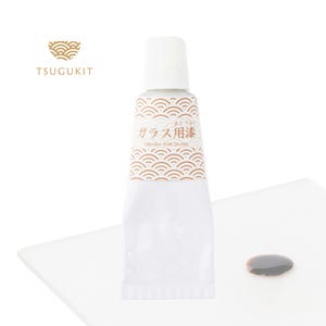 Könnte beinhalten: Eine weiße Tube "Urushi for Glass"-Farbe mit japanischem Text und einem dekorativen Wellenmuster. Neben der Tube befindet sich eine kleine Pfütze dunkelbrauner Farbe auf einer weißen Oberfläche. Der Markenname "TSUGUKIT" ist sichtbar.