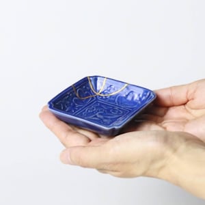 May include: A small, square, cobalt blue ceramic dish with intricate embossed patterns. The dish features gold-colored kintsugi repair lines. The dish is held in two hands against a white background.