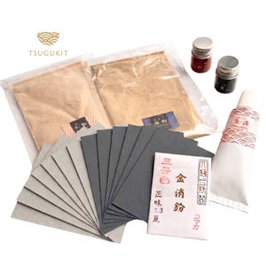 May include: A kintsugi repair kit containing two clear plastic pouches of gold powder, a white tube of lacquer with a red and white patterned label, two small dark jars, and a stack of grey sandpaper sheets with a white paper packet featuring Japanese text.