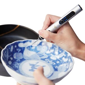 May include: A person is using a silver and blue electric pen tool to decorate a white ceramic bowl with a blue and white floral design.