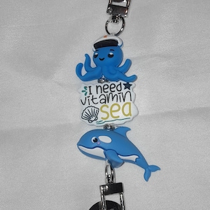 May include: A lanyard charm featuring a blue octopus wearing a white hat, a white rectangular tag with the text I need vitamin sea and a seashell graphic, and a blue orca whale. The charm includes silver-toned metal lobster clasps at both ends.