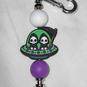 May include: A keychain featuring a green UFO with two skeleton figures in black cloaks. The keychain has a white bead, a purple bead, and a silver spike charm. A silver carabiner clip is attached to the top.