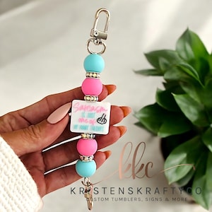 May include: A beaded keychain with a silver clasp, featuring turquoise and pink beads. A white square with the text "Sarcasm is just one of my many talents" is centered. The keychain has a gold-colored charm and a silver clip.